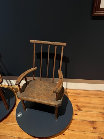 A low, wide-armed antique chair with curved arms on a round museum plinth.